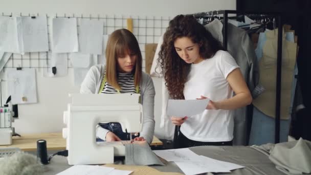 Die professionelle Näherin näht mit der Nähmaschine Stoffe und passt dann die Ausrüstung an, während ihre Kollegin ihre Skizzen zeigt. Atelier mit Schneiderarbeiten im Hintergrund. — Stockvideo