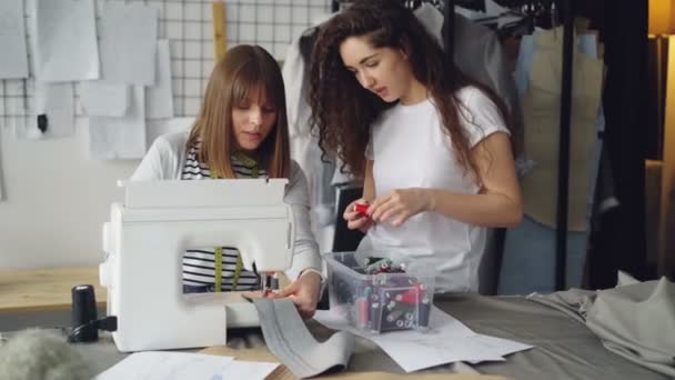 Young woman female designer is working with sewing machine while her partner is looking at stitches and choosing threads from box. Informal friendly atmosphere. — Stock Video