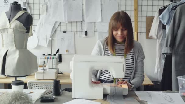 Young attractive dressmaker is working on sewing machine and looking at womens garment sketches in her studio at table. Modern equipment and fashionable garments in background. — Stock Video