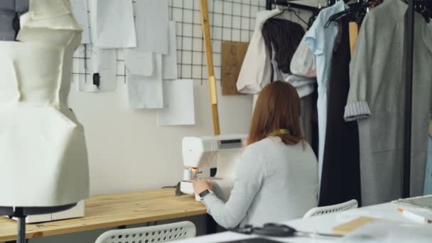 Arkadan görünüşü ile dikiş makinesi studio masa başında oturan genç kadın giyim Tasarımcısı çalışma. Öğeleri, kukla ve kadın Dikiş dikiş elbise görülebilir. — Stok video