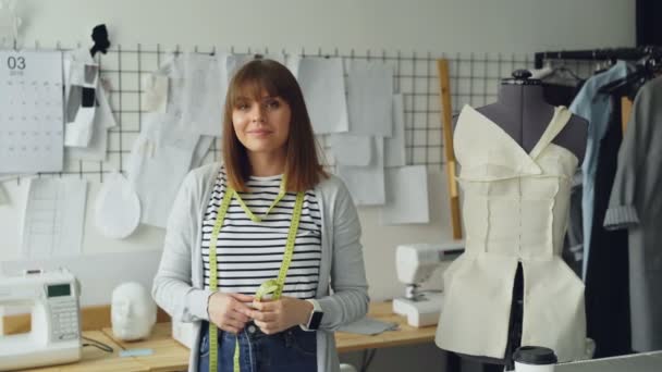 Portret van mooie jonge vrouw naaister permanent in werkplek, ontroerende meetlint en camera te kijken. Pop, kleding, schetsen en naaimachine zijn zichtbaar. — Stockvideo