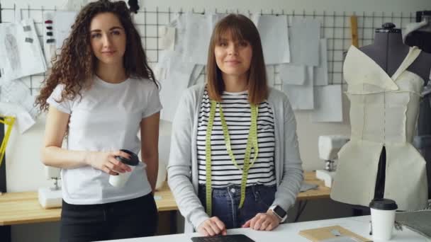 2 女性の服デザイナー起業家ワーク ショップに一緒に立って、カメラ目線の肖像画。一人の女性は、テイクアウトのコーヒーを保持している、他のスタジオの机に触れています。. — ストック動画