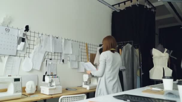 女性服のデザイナーはスタジオのテーブルからスケッチを取ると女性の衣服の他の図面に壁にそれらを置きます。創造的な思考の概念. — ストック動画