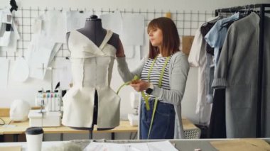 Genç kadın terzisi ölçme giyim tutturulmuş kukla ölçü-bant ile terzilik için. Konsantre ve düşünceli bir kadındır. Stüdyo hafif, birçok dikiş araç ve öğeleri ile modern.