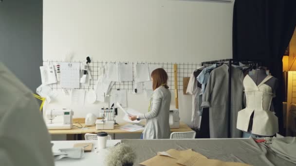 Female fashion designer is choosing and hanging clothing sketches to wall for her newest collection. Light fabrics, clothes hanging and sewing items are visible. — Stock Video