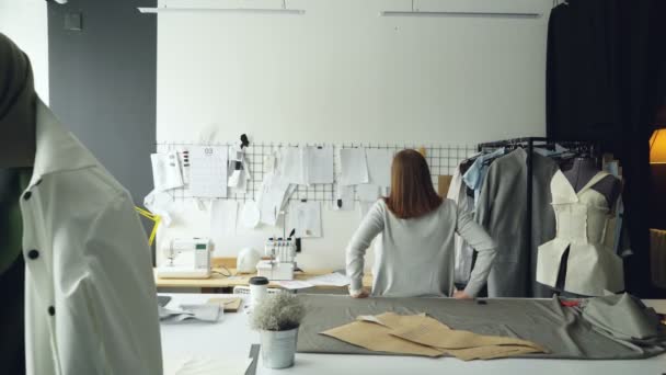 Die junge Designerin steht mit dem Rücken zur Kamera und betrachtet Zeichnungen, die an der Wand hängen. Frau denkt über neue Kleidersammlung nach. — Stockvideo