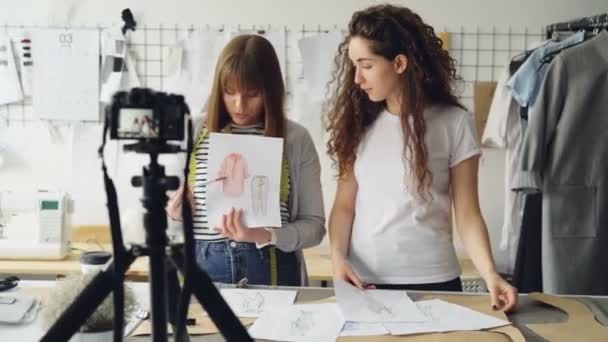 Bloggers de moda de mujeres jóvenes grabando video blog sobre señoritas en cámara y hablando con seguidores en el estudio moderno. Muchos bocetos de prendas son visibles . — Vídeo de stock