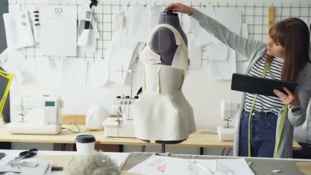 Hermoso sastre está convirtiendo maniquí de sastrería para comprobar la ropa fijada y trabajar con la tableta para introducir información. Uso de tecnologías modernas en el concepto de fabricación de ropa . — Vídeos de Stock