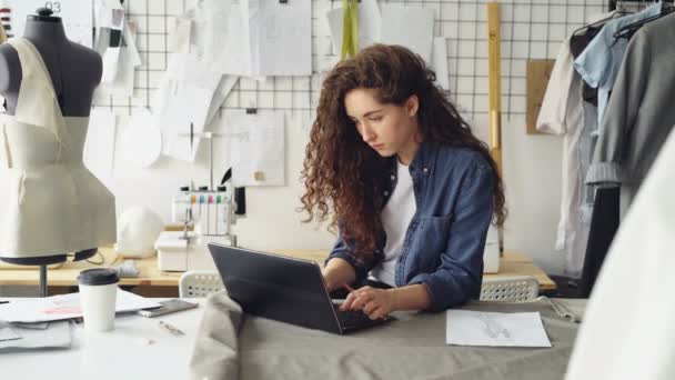 Costurera autónoma está trabajando con el ordenador portátil sentado en la mesa en el taller de luz moderna. Mujer está escribiendo y viendo scree, a continuación, dibujar boceto de la prenda . — Vídeos de Stock