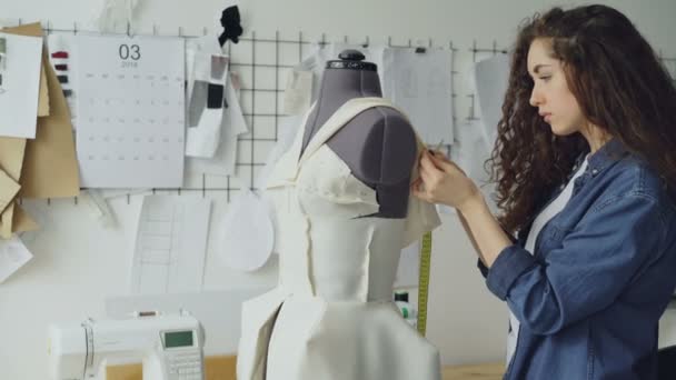 若い女性のテーラーを服に仕立て縫製ピンをダミーにしてメジャー テープを測定調整します。レディース衣類項目を仕立ての壁にスケッチが表示されます。. — ストック動画