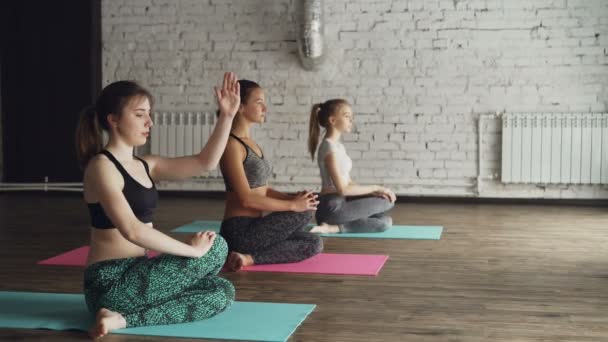 Instructor de yoga está demostrando brazos giro en la cara de vaca posan Gomukhasana mientras los estudiantes escuchan y prueban esta posición. Vista lateral de las mujeres jóvenes es un estudio con paredes blancas . — Vídeos de Stock