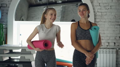 Birlikte spor salonunda yürüyüş ve duygusal konuşuyor cheeful genç kadın Dolly atış. Kadınlar yoga paspaslar taşıyorsun, parlak spor malzemeleri görülür.
