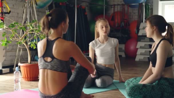 Yoga students are sitting on mats and talking while waiting for morning practice in large light gym. Fitness balls, step aerobic platforms and other sports equipment is visible. — Stock Video