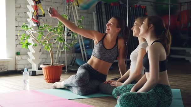 Yoga students are taking selfie with smartphone while sitting together on bright mat in yoga studio. Women are posing, smiling and laughing. Wellness fun concept. — Stock Video