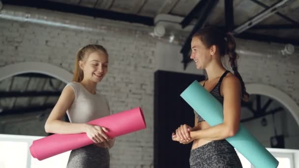 Chicas atractivas están hablando y riendo en el gran centro deportivo moderno. Las mujeres sostienen coloridas esteras de yoga y usan ropa deportiva de moda. . — Vídeos de Stock