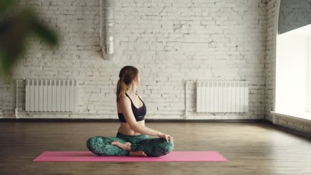 La joven con ropa deportiva de moda está haciendo giros corporales sentada en posición de loto en la alfombra dentro del estudio de yoga. Relajante concepto de práctica personal . — Vídeos de Stock