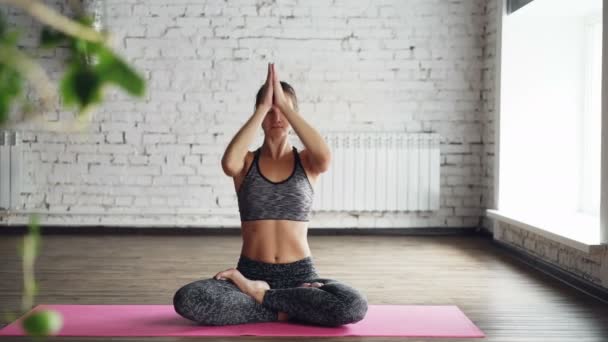 Mujer atractiva joven está teniendo clase de yoga individual. Ella está sentada sobre una alfombra en pose de loto, levantando las manos y luego bajándolas en namaste, poniéndolas de rodillas y relajándose. . — Vídeos de Stock