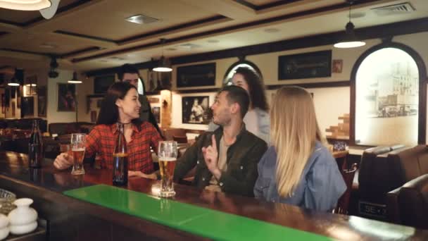 Junge Leute unterhalten sich an der Theke bei Bier, wenn ihnen ein paar ihrer Freunde entgegenkommen. Männer und Frauen begrüßen sich und teilen Neuigkeiten. — Stockvideo