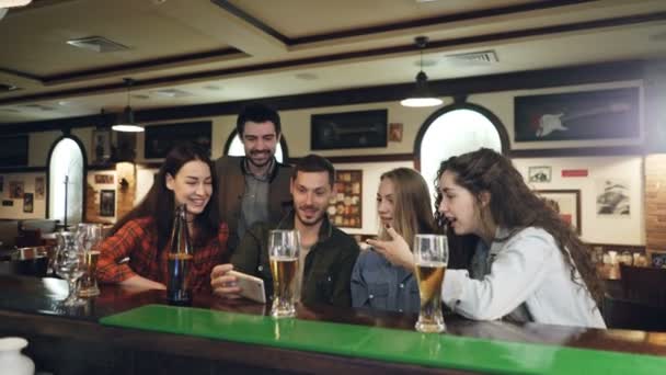 Красивий бородатий хлопець показує відео на смартфоні своїм друзям, розслабляючись у барі разом. Молоді люди обговорюють і сміються, вказуючи на екран . — стокове відео