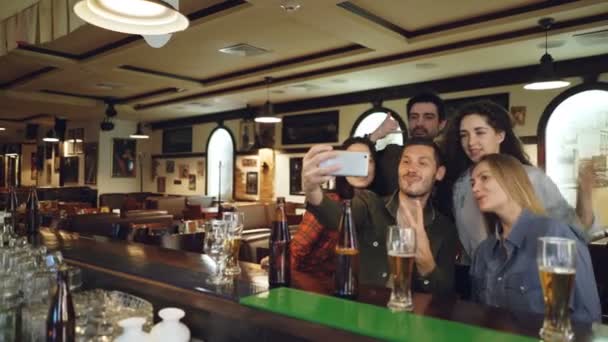 Friends are taking selfie with smartphone in bar. Young people are posing, laughing and talking. Beer bottles and glasses in foreground — Stock Video