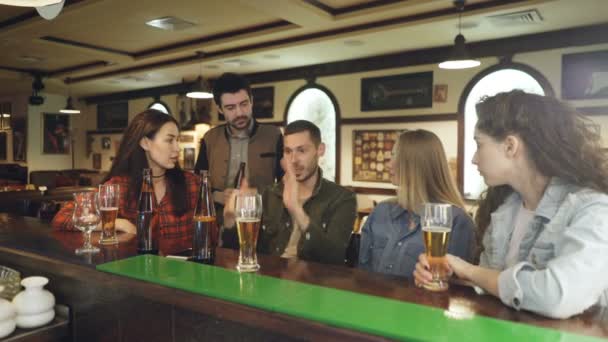 Bonito jovem barbudo está conversando com seus amigos e gesticulando emocionalmente enquanto está sentado no pub local no balcão. Os seus amigos estão a ouvi-lo com atenção. . — Vídeo de Stock