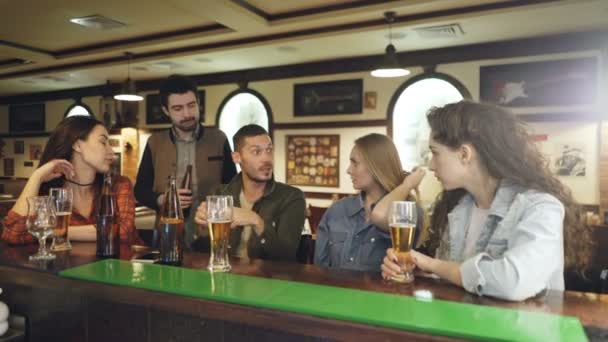 Jovem barbudo de cabelos escuros está contando uma história e gesticulando emocionalmente enquanto se encontra com seus amigos no pub no balcão. Os seus amigos estão a ouvi-lo com atenção. . — Vídeo de Stock