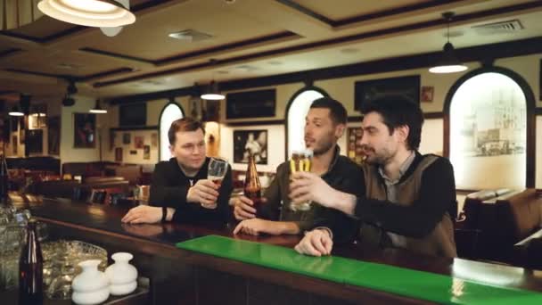 Tres jóvenes con ropa casual están bebiendo cerveza en un elegante bar deportivo, colgando vasos y botellas y hablando mientras están sentados en el mostrador. Chicos teniendo concepto de salida nocturna . — Vídeos de Stock