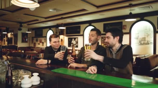 Three young men in casual clothes are drinking beer in fancy sports bar, clanging glasses and bottles and talking while sitting at counter. Guys having night out concept.