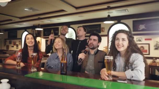 Jovens amigos felizes estão cantando karaoke enquanto bebem cerveja no bar moderno. Eles estão desfrutando de bebidas, música e boa companhia. Companheiros ter conceito divertido . — Vídeo de Stock