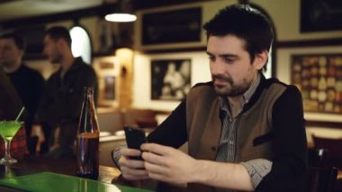 Genç erkek öğrenci şişe bira ile süslü barda oturan akıllı telefon kullanıyor. O ekran dokunaklı ve gülümseyerek. İletişim kavramı modern yolları.