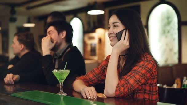 Jovem morena atraente está chamando seu namorado enquanto está sentada sozinha no balcão no bar moderno . — Vídeo de Stock