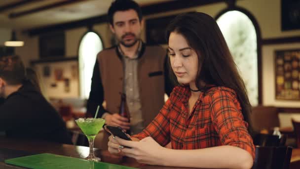 ビール瓶を持つハンサムな男が来ると彼女に話しているとき、若い女性がバーのカウンターで、スマート フォンを使用しています。ガラスをチリンとして社交. — ストック動画