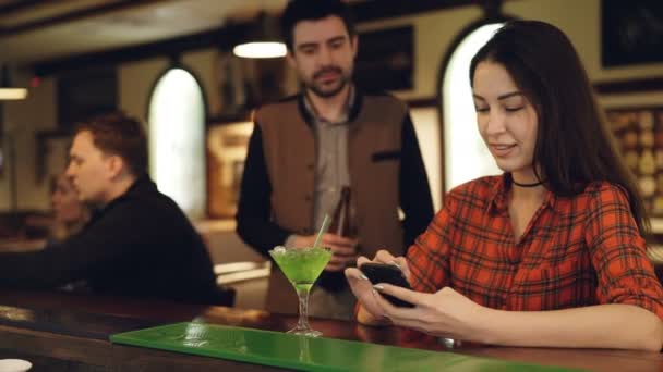 Atractiva morena está revisando el teléfono inteligente en el mostrador del bar cuando el joven se está familiarizando con ella. Los jóvenes están charlando, sonriendo y tintineando gafas . — Vídeo de stock