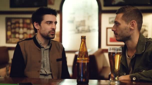 Due vecchi amici stanno conversando al pub davanti alla birra. Vetro e bottiglia in primo piano, tavoli e immagini su pareti in sottofondo . — Video Stock
