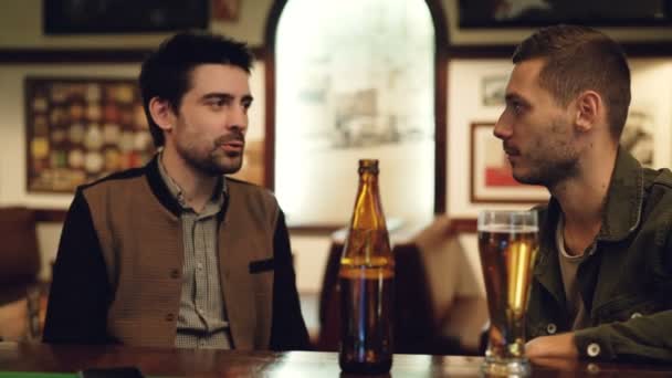 Dos viejos amigos están conversando en el pub sobre la cerveza. Vidrio y botella en primer plano, mesas e imágenes en las paredes en el fondo . — Vídeo de stock