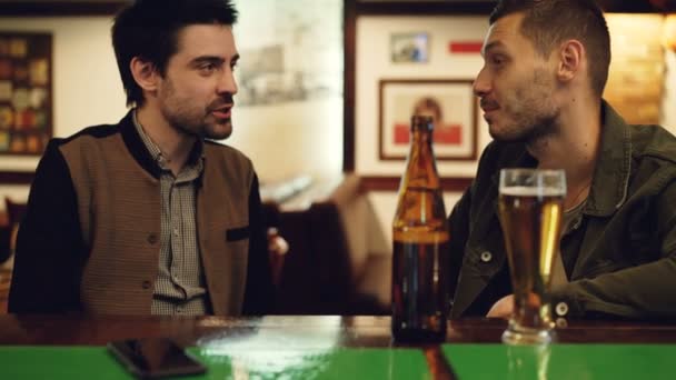 Los alegres amigos varones están charlando y bebiendo cerveza en un bonito pub. Los hombres guapos están hablando, golpeando las gafas y riendo mientras descansan en el bar — Vídeos de Stock