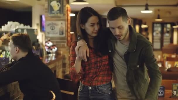 Dolly shot of young woman helping her drunk boyfriend leave bar. Handsome bearded man is holding beer bottle and telling something to his girlfriend. — Stock Video