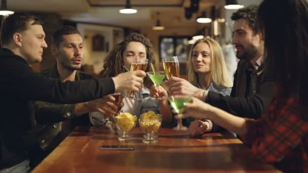 Des jeunes gens joyeux mangent, boivent, bavardent et rient à table dans un café moderne. Les copains portent des vêtements décontractés. Snacks et verres au premier plan . — Video