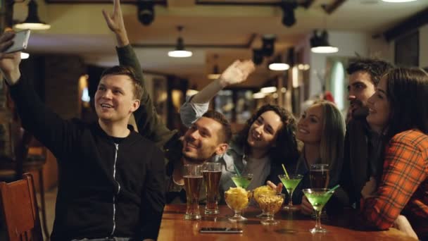 El joven bloguero está grabando video con un smartphone sentado en la mesa en el bar con sus amigos. Los jóvenes posan, saludan con las manos y ríen. Blogging para el concepto de instagram . — Vídeo de stock