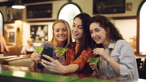Attractive young women are taking selfie with cocktails in bar. Cheerful girls are posing, laughing and clanging glasses. Taking pictures for instagram account concept. — Stock Video