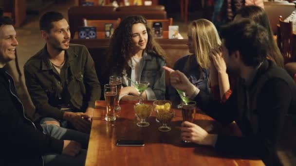 Alegre hombres y mujeres jóvenes están celebrando evento importante en el bar agradable. Son vasos colgantes, tostadas y bebidas. Amigos divirtiéndose en concepto de fin de semana . — Vídeo de stock