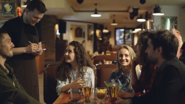 Los jóvenes están hablando con el camarero haciendo orden en la cafetería mientras el camarero escucha a los clientes y escribe en bloc de notas con bolígrafo. Comer y beber fuera del concepto . — Vídeo de stock