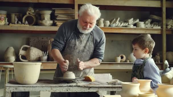 Caring senior grandfather is showing young grandson how to work with clay on throwing-wheel in small workshop. Pottery, family hobby and handicraft concept. — Stock Video