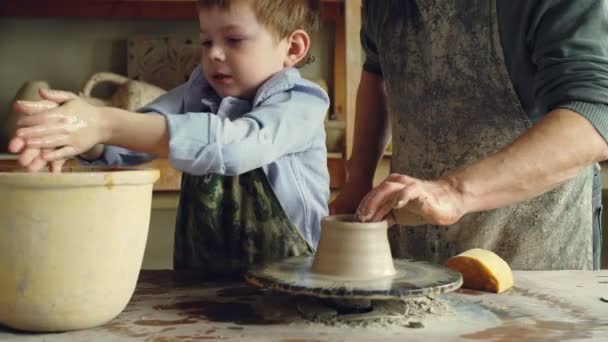 Lindo niño está aprendiendo cerámica en el taller tradicional junto con el abuelo mayor. Él está mojando las manos, formando un pedazo de arcilla en la rueda de lanzamiento . — Vídeos de Stock