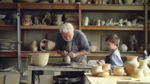 Vedoucí keramik je formovat hlíny na spinning házení kola, zatímco jeho vnuka je sledovat a učení. Sdílení zkušeností, rodinné tradice a ruční koncepce. — Stock video