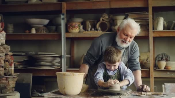Concentrated child is forming pot from clay on potters wheel under guidance of his experienced grandfather. Ceramic pots, vases and figures are in background. — Stock Video