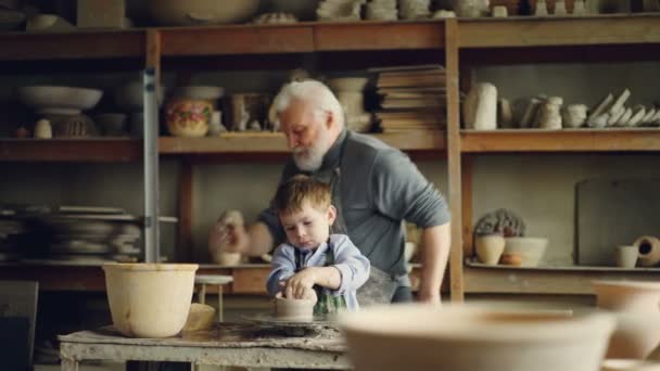 Elderly man experienced potter is teaching little boy how to work with clay on potters wheel. Sharing experience, family tradition and modern pottery concept. — Stock Video