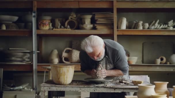 Hardworking grey-haired man is working with clay on potters wheel, shaping piece of loam. Beautiful ceramic utensils, handmade pots and vases on shelves are visible. — Stock Video