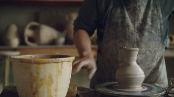 Elderly craftsman is creating ceramic jar on potters wheel using professional tools. Workplace with handmade pots, beautiful vases and figures in background. — Stock Video