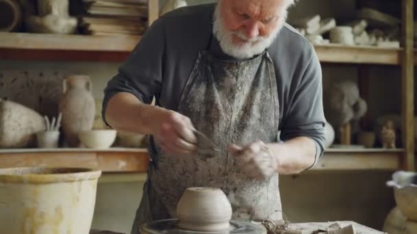 Ceramista senior es suavizar la parte inferior de la mitad de acabado de olla de cerámica en la rueda de alfareros girando en el lugar de trabajo. Creación de utensilios de cocina tradicionales y concepto de cerámica convencional . — Vídeos de Stock